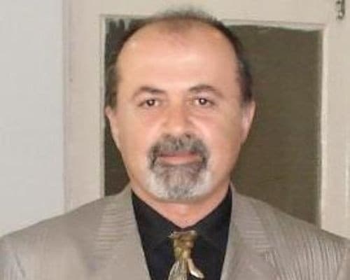 This image shows a man wearing a well-tailored suit. He is standing with his hands in his pockets, looking off into the distance. He has a confident and determined expression on his face. His suit is composed of a navy blue jacket and trousers, with a white shirt and a thin black tie. He is wearing black leather dress shoes. His eyes are a warm brown color, and his hair is neatly combed back. His posture is straight and his chin is slightly raised in a proud and dignified manner. He looks like a successful businessman, ready to take on any challenge that comes his way.