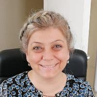 Omuz hizasında koyu renk saçlı bir kadın doğrudan kameraya bakıyor ve gülümsüyor. Dostça bir ifadesi var ve gözleri parlak ve uyanık. Yanakları hafifçe pembe ve dudakları hafifçe ayrık. Üzerinde v yakalı beyaz bir tişört ve mavi bir kot pantolon var. Ellerini kalçalarına koymuş, rahat ve kendinden emin bir poz veriyor. Çevresinden memnun ve mutlu görünüyor.