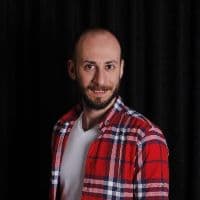 A young man is standing in front of a plain background wearing a bright red and black plaid shirt. He has short dark hair and is smiling slightly, with his arms crossed. He is wearing black jeans and black shoes. His posture is confident and relaxed. He appears to be in his late twenties or early thirties, with a youthful, healthy complexion. He is standing in a three-quarters pose, looking directly at the camera. He appears to be enjoying the moment, with a friendly expression on his face.