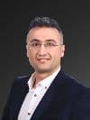 Özel dikim siyah takım elbiseli yakışıklı bir adam gri bir arka planın önünde kendinden emin bir şekilde duruyor. Kısa, koyu renk saçları ve hafif bir sakalı var ve gözleri uzaklara bakıyor. Duruşu rahat, elleri rahatça ceplerinde ve dudaklarında hafif bir gülümseme var. Zarif ve sofistike görünüyor ve hayatın sunacağı her şeyi üstlenmeye hazır. Beyaz gömleği ve siyah kravatıyla kıyafetini tamamlıyor ve görünümüne sofistike bir hava katıyor.