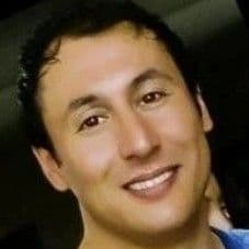 Parlak bir gülümsemeye sahip yakışıklı bir adam doğrudan kameraya bakıyor. Koyu kahverengi gözleri, kısa koyu kahverengi saçları ve güçlü bir çene hattı var. Beyaz bir tişört giyiyor ve sol kulağında küçük bir küpe var. Yüz ifadesi rahat ve memnun. Çenesinde hafif bir kirli sakal ve yanaklarında birkaç çil var. Yirmili yaşlarının ortalarında görünüyor ve sağlıklı ve mutlu görünüyor.