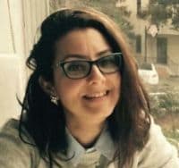 Gözlük takan ve genişçe gülümseyen genç bir kadın. Orta uzunlukta kahverengi saçları var ve gözleri parlak ve ışıltılı. Gülümsemesi geniş ve doğrudan kameraya bakıyor. Pembe yanakları ve dostça bir ifadesi var. Gözlükleri kahverenginin açık bir tonunda ve yüzünde hafifçe orantısız duruyor. Üzerinde gri bir gömlek ve boynunda siyah bir eşarp var. Rahat ve mutlu görünüyor.