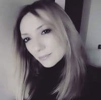 Omuz hizasında kahverengi saçları olan genç bir kadın, yüzünün önünde tuttuğu beyaz bir akıllı telefonla selfie çekiyor. Parlak, neşeli bir gülümsemesi var ve gözleri tamamen açık. Sağ eli telefonu desteklerken sol eli yüzünün yan tarafında duruyor. Mavi beyaz çizgili bir tişört ve bir çift kot şort giymektedir. Saçları hafifçe dağılmış ve rahat, kaygısız bir pozu var. Gri bir duvarın önünde duruyor ve etrafı parlak, güneşli bir atmosferle çevrili. Bu neşeli an, anı yaşamanın sevincini yakalıyor.