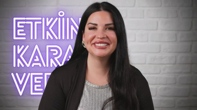 Bir kadın yüzünde parlak bir gülümsemeyle ayakta duruyor, gözleri kapalı ve başı hafifçe yana eğik. Koyu renkli bir bluz giymiş ve saçları yüzünün ve boynunun etrafına dökülüyor. Dudakları hafifçe ayrılmış ve dişleri görünüyor. Üzerinde görünmeyen bir tabela bulunan bir duvarın önünde durmaktadır. Çenesi yukarı doğru eğiktir ve yakın çekimde boynu görünür. Fotoğraf için gülümsüyor, ifadesi mutluluk ve memnuniyet hissi veriyor.