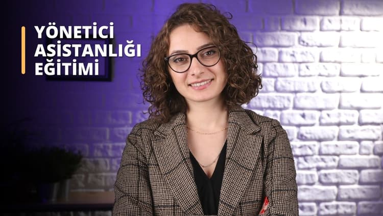 Açık kahverengi ceket giyen ve yuvarlak, siyah çerçeveli gözlük takan bir kadın kapalı bir ortamda duruyor. Yüzünde hoş bir ifade var, dudakları hafifçe gülümseme şeklinde kıvrılmış. Saçları kısa ve kıvırcıktır ve boynuna altın bir kolye takmaktadır. Arka plan, üzerinde beyaz yazılar olan mor bir duvardır. Arka planda bir bitkinin bulanık bir görüntüsü var.