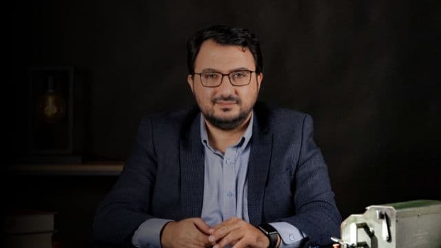 Bu, koyu mavi takım elbise giyen bir adamın fotoğrafı. Arkasında beyaz bir duvar olan parlak ışıklı bir odada durmaktadır. Adamın kısa siyah saçları var ve yüzünde ciddi bir ifadeyle önüne bakıyor. Beyaz bir elbise gömleği, mavi çizgili bir kravat ve koyu mavi bir takım elbise ceketi giymektedir. Ayrıca siyah elbise pantolonu ve siyah elbise ayakkabıları giymektedir. Elleri yanındadır ve sol bileğinde bir saat takılıdır. Adam kendinden emin ve sakin görünüyor.