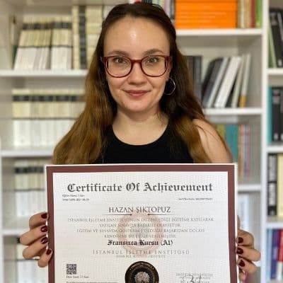 Ön planda bir kadın var. Elinde İstanbul işletme Enstütüsüne ait bir sertifika var. Başarılı olduğu için sertifika aldığını anlatıyor. Arka planda kitaplık ve bir çok kitap yer oluyor. Portre fotoğrafına benziyor. Profesyonel fotoğraf makinesiyle çekilmiş.