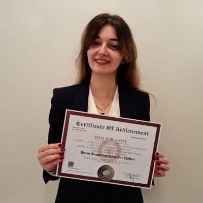 This photo displays a smiling woman holding a certificate in her hands. She has long dark hair that is pulled back into a neat bun, and wears a white blouse with a black skirt. The certificate is white and green with a gold seal and text on the front. She has a look of pride and accomplishment on her face, likely due to receiving the certificate. The background is a neutral off-white wall, with a few other certificates hung up behind her. This image could symbolize the feelings of success and achievement that come with a hard-earned accomplishment.