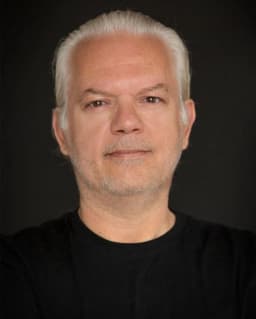 Bu görüntüdeki adam beyaz saçlı ve düz siyah bir gömlek giyiyor. Yirmili yaşlarının ortalarında olduğu anlaşılıyor ve temiz traşlı bir yüzü var. İfadesi nötr ve gözleri kapalı. Resmin arka planı çoğunlukla aydınlık, sağ alt köşede biraz koyu gölgeler var. Büyük bir pencerenin önünde duruyor ve içeriye ışık giriyor. Duruşu kendinden emin ve elleri ceplerinde. Düşüncelere dalmış gibi görünüyor ve varlığı sakinleştirici ve huzur verici. Beyaz saçları gömleğinin siyahına karşı öne çıkıyor ve giysileri sade ve zamansız. Sakinlik hissi veren huzurlu ve dingin bir görüntü.