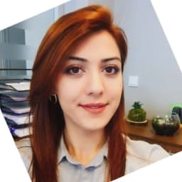 Genç bir kadın beyaz bir arka planla selfie çekiyor. Bir duvarın önünde duruyor ve kameraya bakarken yüzünde parlak, kendinden emin bir gülümseme var. Uzun, koyu renk saçları gevşek bir topuzla geriye toplanmış ve üzerinde siyah beyaz desenli gri bir tişört var. Ellerini uzatmış, biri telefonu yüzüne doğru tutarken diğeri 'V' şeklinde kaldırmış. Işık yüzünden yansıyarak yüz hatlarını vurguluyor. Mutlu ve memnun görünüyor, anın tadını çıkarıyor ve onu kendisi için yakalıyor.