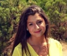 Kadın ışıl ışıl gülümsüyor, yüzü fotoğraf makinesinin flaşının sıcak ışıltısıyla aydınlanıyor. Gözleri kapalı, dudakları neşeli bir ifadeyle hafifçe ayrılmış. Saçları geriye doğru toplanmış, yüzünü çerçeveliyor ve gülümsemesini vurguluyor. Açık pembe bir gömlek ve koyu mavi bir kot pantolon giymiş, kıyafeti neşeli tavrını tamamlıyor. Rahat ve memnun görünüyor, elleri yanında duruyor. Bakışları kameradan hafifçe uzağa yönelmiş, bu da huzur ve memnuniyet hissi uyandırıyor. Duruşu kendinden emin ve gururlu, gülümsemesi neşe ve memnuniyet hissi yayıyor.