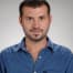 Parlak mavi gömlekli bir adam düz bir arka planın önünde duruyor. Kısa siyah saçları ve temiz traşlı bir yüzü vardır. Elleri arkasında, kameradan uzağa bakmaktadır. Vücudu hafifçe yana dönüktür ve izleyiciye neredeyse profilden bir görünüm verir. Üzerinde beyaz cepli açık mavi yakalı bir gömlek ve koyu mavi bir kot pantolon var. Duruşu rahat ve gevşek bir tavır sergilediğini gösteriyor. İfadesi memnuniyet ve huzur dolu. Gömleğinin mavisi nötr arka planla güzel bir tezat oluşturarak görsel açıdan hoş bir görüntü yaratıyor.