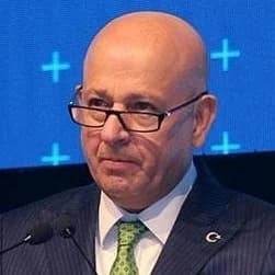 Takım elbiseli adam yüzünde ciddi bir ifadeyle dimdik ayakta duruyor. Açık mavi takım elbisesi tam oturmuş ve aynı derecede net beyaz bir gömlek ve koyu mavi kravatla tamamlanmış. Saçları düzgünce geriye taranmış ve gözleri uzaktaki bir şeye odaklanmış. Parlatılmış siyah deri elbise ayakkabıları giyiyor. Elleri önünde birbirine kenetlenmiş ve bir şey bekliyor gibi görünüyor. Duruşu kendinden emin ve ağırbaşlı, profesyonellik ve yetkinlik havası taşıyor. Önüne çıkan her türlü zorluğun üstesinden gelmeye hazır görünüyor.