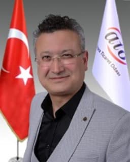 İyi giyimli, gözlüklü bir adam aydınlık bir odada durmaktadır. Üzerinde siyah bir takım elbise, altında beyaz bir gömlek ve kalın çerçeveli bir gözlük var. Saçları düzgünce şekillendirilmiş ve nötr bir ifadeyle dümdüz karşıya bakıyor. Elleri hafifçe arkada, ayakta duruyor ve duruşu kendinden emin ve sakin. Otuzlu yaşlarının ortalarında ve orta yapılı görünüyor. Bakışları odaklanmış ve yoğun, yüz hatları keskin ve iyi tanımlanmış. Kendisi saygın bir profesyonel ve bu görüntü bir güven ve ciddiyet hissi veriyor.