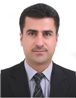 Şık giyimli bir adam, üç parçalı takım elbisesi ve kravatıyla kendinden emin bir şekilde duruyor. Siyah bir takım elbise, beyaz bir gömlek ve kırmızı bir kravat giymiştir. Adamın düzgünce taranmış siyah saçları ve temiz traşlı bir yüzü vardır. Elleri önünde kenetlenmiş ve yüzünde hafif bir gülümsemeyle ileriye bakıyor. Solunda bir pencere bulunan gri bir duvarın önünde duruyor ve dışarıdan gelen ışık siluetine gölge düşürüyor. Duruşu güç, güven ve gurur duygusu yayıyor.