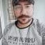 Bıyıklı ve sakallı genç bir adam selfie çekiyor. Telefonunu önünde tutuyor ve kararlı bir ifadeyle objektife bakıyor. Yüzündeki kıllar düzgün, kalın bir bıyık ve dolgun, kalın bir sakal şeklinde bakımlı. Siyah bir tişört ve koyu renk bir kot pantolon giymiş. Arka plan net değil ama kadrajda bir oturma odasının izleri var. Bu selfie'yi çekerken gözleri odaklanmış ve konsantre olmuş durumda. Anın tadını çıkarıyor gibi görünüyor ve yüzü sıcak, mutlu bir ifadeyle aydınlanıyor.