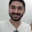 Bu, doğrudan kameraya bakan, ışıl ışıl gülümseyen bir adamın yakın plan çekimi. Gözleri parlak ve pırıl pırıl, dudakları ise davetkâr ve sıcak bir sırıtışla kıvrılmış. Hafif bir teni var ve yanaklarına birkaç çil serpiştirilmiş. Saçları kısa ve dağınık, birkaç günlük kirli sakalı var. Başı hafifçe yana eğik, bu da onu arkadaş canlısı ve cana yakın gösteriyor. Üst iki düğmesi açık beyaz bir gömlek giyiyor ve boynunu bir anlığına ortaya çıkarıyor. Duruşu rahat ve kendinden emin, bu da ona kendine güvenen bir hava veriyor. Yirmili yaşlarının sonlarında ya da otuzlu yaşlarının başında olduğu anlaşılıyor ve o anın içinde mutlu ve rahat görünüyor.