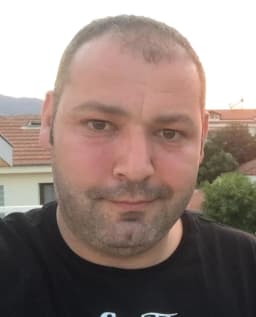 Siyah saçlı ve açık mavi tişörtlü bir adam beyaz bir duvarın önünde duruyor ve sağ elinde bir akıllı telefon tutuyor. Sol elini önüne doğru uzatmış ve telefonunun ön kamerasına bakarak selfie çekiyor. Yüzünde parlak bir gülümseme var ve anın tadını çıkarıyor gibi görünüyor. Işık yumuşak ve görüntüdeki renkler canlı. Adam dik bir duruş sergiliyor ve kameraya kendinden emin bir şekilde bakıyor.