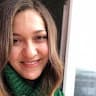 Uzun, siyah saçlı ve parlak bir gülümsemeye sahip genç bir kadın kameraya bakıyor. Yüzü neşe dolu ve gözleri ışıl ışıl. Üzerinde pembe beyaz çizgili bir gömlek var ve dudakları neşeli bir ifadeyle hafifçe aralanmış. Elleri önünde kenetlenmiş ve kolları hafifçe bükülmüş. Açık gri bir duvarın önünde duruyor ve ışık yüzünde parlayarak yüz hatlarını belirginleştiriyor. Rahat ve kendinden emin görünüyor ve tüm vücudu mutluluk yayıyor. Yüzü saçlarıyla çerçevelenmiş ve güzel gülümsemesi görüntünün odak noktasını oluşturuyor.