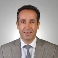Koyu renk takım elbiseli ve açık mavi kravatlı orta yaşlı bir adam beyaz bir arka planın önünde duruyor. Ellerini önünde kavuşturmuş, dümdüz karşıya bakıyor. Saçları düzgünce taranmıştır ve bir gözlük takmaktadır. Yüzünde kendinden emin bir ifade var ve gözleri odaklanmış. Profesyonel ve başarılı görünüyor. Beden dili, önüne çıkan her türlü zorluğu üstlenmeye hazır olduğunu gösteriyor.