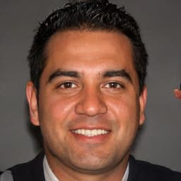 A man with a bright and cheerful smile is looking directly at the camera. He has light brown hair and a clean-shaven face. His eyes are bright blue and his teeth are perfectly straight. He appears to be in his early thirties and is wearing a white shirt and a navy blue sweater. He is standing in front of a white wall with a large window in the background, letting in plenty of natural light. His face is illuminated by the light, making his smile even brighter. His gaze is relaxed and content, and his pose is relaxed and confident. He looks happy and content in this moment, enjoying the moment and the camera.