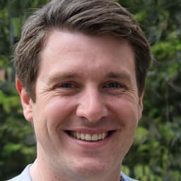 A man is standing in front of the camera, looking directly at the lens with a wide smile on his face. He is wearing a white collared shirt and dark pants. His hair is short and neatly combed back. His eyes are bright and his cheeks are rosy. His hands are in his pockets and his feet are slightly apart. He is standing in a natural, relaxed pose. His expression conveys a feeling of joy and contentment. He appears to be enjoying the moment, and is ready to capture the memory with a picture.