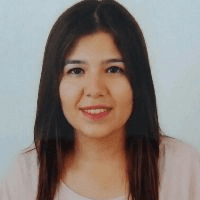 Omuz hizasında kumral saçları olan bir kadın yüzünde kocaman, ışıl ışıl bir gülümsemeyle ayakta duruyor. Üzerinde beyaz yakalı ve çiçek desenli pembe bir bluz var ve saçları rüzgârda hafifçe dalgalanıyor. Dudakları neşeli bir ifadeyle kıvrılmış, gözleri parlak ve mutlu. Ellerini kalçalarına koymuş ve soluk mavi bir arka planın önünde duruyor. Uzun bukleleri sırtından aşağı dökülüyor ve ışıkta parlıyor. Memnun ve neşeli görünüyor, güven ve pozitiflik duygusu yayıyor.