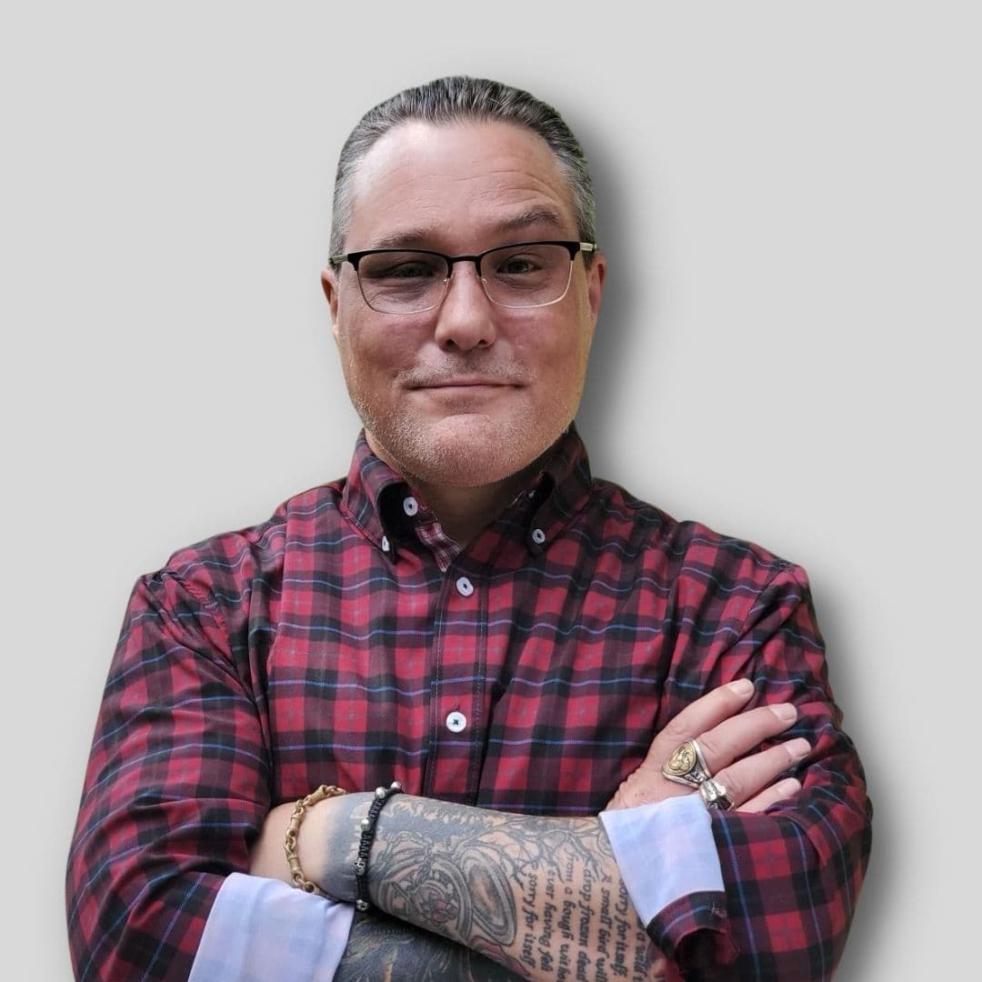 A man with short brown hair stands looking at the camera with his arms stretched out. His arms are covered in intricate black and white tattoos that depict a variety of symbols and shapes. He wears a black t-shirt and blue jeans, and his dark eyes appear to be looking off into the distance. His facial expression is serious yet thoughtful, conveying a deep sense of concentration and contemplation. His strong jawline and chiseled features give him an air of confidence and conviction. He stands proudly, a silent testament to the artwork displayed on his body.