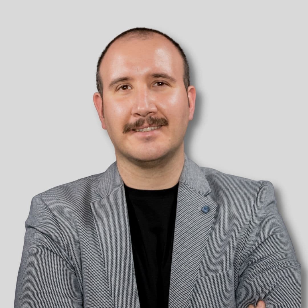 The man is wearing a smart, dark grey suit. He has a neatly trimmed moustache, and his hair is combed neatly to the side. His eyes are focused forward as if looking into the distance. He has a confident and serious expression on his face. He is standing straight with his hands on his hips in a proud pose. He looks confident and determined. His posture exudes a sense of strength and control.