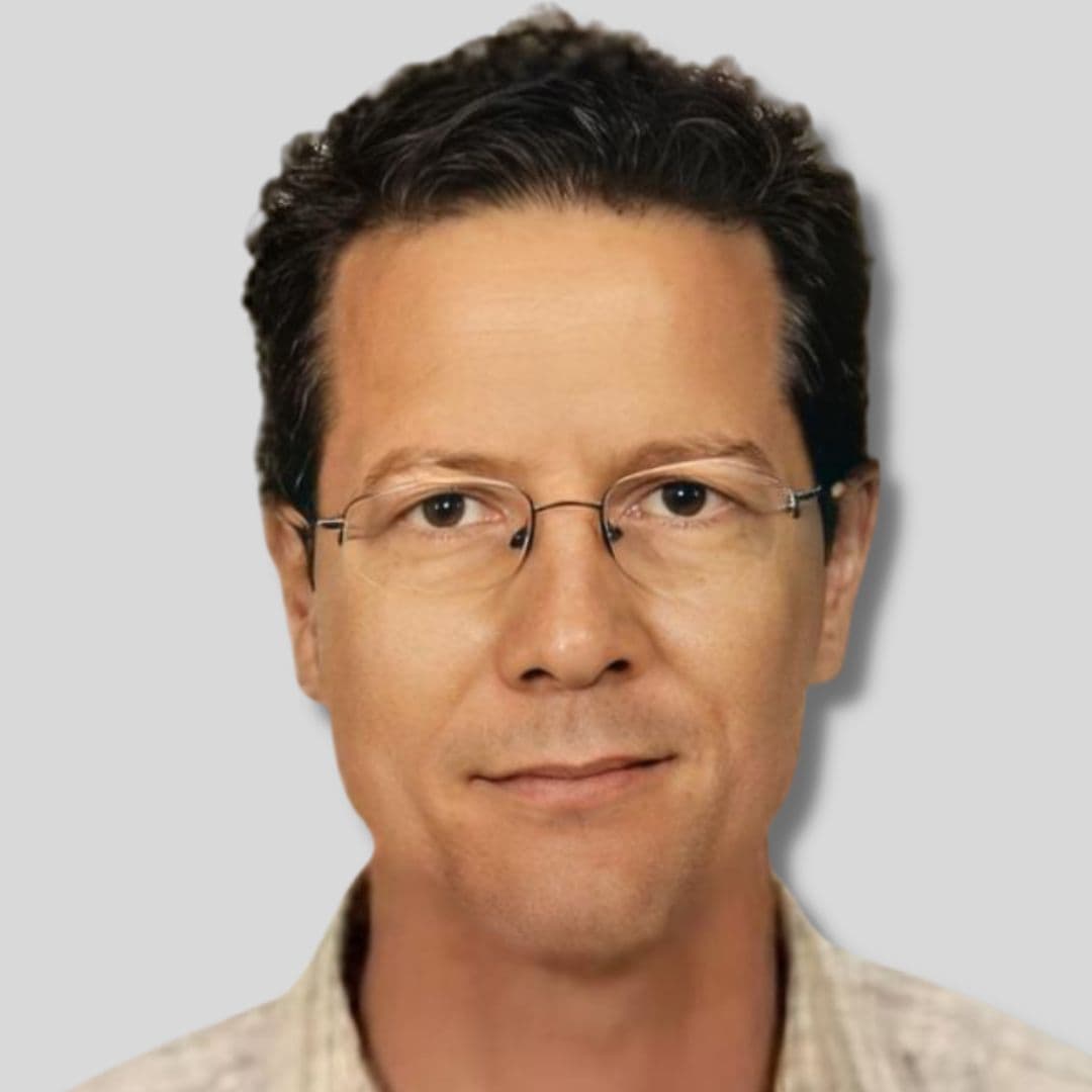 A middle-aged man wearing a white, collared button-up shirt and glasses stares directly at the camera. He has dark, short hair, a strong jawline, and a confident expression. His shirt is unbuttoned at the top, and the sleeves are rolled up to his elbows. He has a light stubble on his chin and cheeks. He has a broad forehead and his eyebrows are slightly raised. His glasses are black and rectangular, and the frames are slightly oversized. Overall, he looks smart and competent.