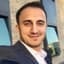 Yüzünde geniş bir gülümseme olan genç bir adam kameranın önünde duruyor. Kısa, siyah saçları ve parlak, mavi gözleri var. Beyaz bir tişört ve kahverengi kemerli siyah bir kot pantolon giyiyor. Kolları iki yanında ve başı hafifçe sola doğru eğik. Rahat bir duruşu var ve anın tadını çıkarıyor gibi görünüyor. Gülümsemesi sıcak ve davetkâr, gözleri ise neşe dolu. Dünyayı ele geçirmeye hazır görünüyor.