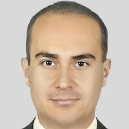 Kısa, siyah saçlı bir adam resmi siyah bir takım elbise giymiş ve mavi kravat takmış. İyi aydınlatılmış bir odada ayakta duruyor ve ciddi bir ifadeyle önüne bakıyor. Ellerini önünde kavuşturmuş ve belinde kahverengi deri bir kemer var. Duruşu kendinden emin ve bakışları odaklanmış. Otuzlu yaşlarının ortalarında görünüyor ve kıyafetleri düzgün ve şık. Kare bir çene çizgisi ve temiz traşlı bir yüzü var. Gözleri kahverengi ve kaşları belirgin. Güçlü ve kararlı bir görünümü var.