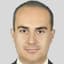 Kısa, siyah saçlı bir adam resmi siyah bir takım elbise giymiş ve mavi kravat takmış. İyi aydınlatılmış bir odada ayakta duruyor ve ciddi bir ifadeyle önüne bakıyor. Ellerini önünde kavuşturmuş ve belinde kahverengi deri bir kemer var. Duruşu kendinden emin ve bakışları odaklanmış. Otuzlu yaşlarının ortalarında görünüyor ve kıyafetleri düzgün ve şık. Kare bir çene çizgisi ve temiz traşlı bir yüzü var. Gözleri kahverengi ve kaşları belirgin. Güçlü ve kararlı bir görünümü var.
