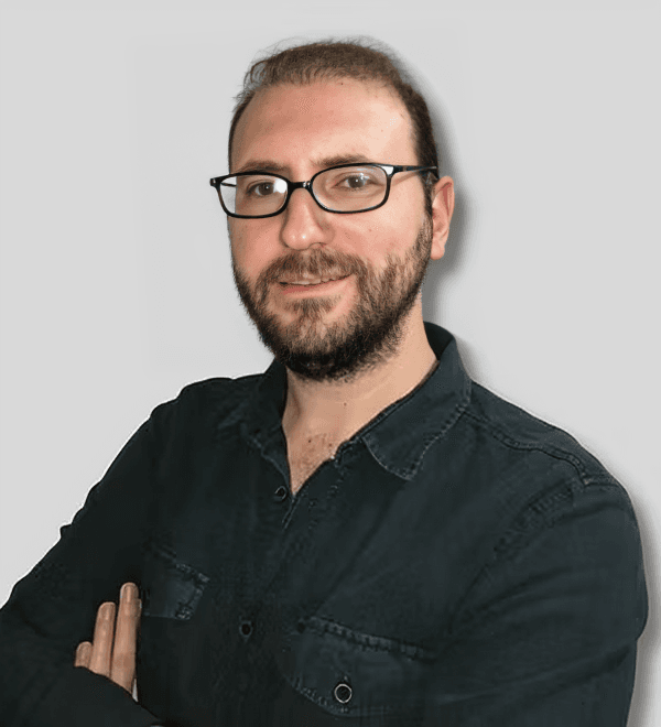 A middle-aged man wearing glasses and a full beard is facing forward with a serious expression on his face. He is wearing a gray, long-sleeve shirt with a black tie and is standing in a white-walled room with a light brown floor. His hair is dark brown and combed back, and his eyes are a brownish-green color. His glasses appear to be square-framed and have lenses that are slightly tinted. His facial features, such as the shape of his nose and his lips, are visible. He looks calm and composed, standing tall and proud.