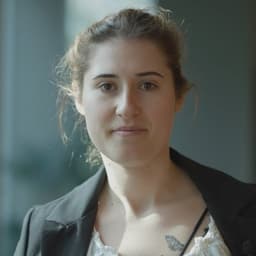Siyah ceket giyen bir kadın kameraya bakıyor ve yeni edindiği sertifika hakkında röportaj veriyor.