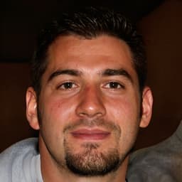 Resimdeki adamın dolgun, koyu renk bir keçi sakalı ve sakalı var. Açık renkli bir gömlek giyiyor ve yüzünde ciddi bir ifade var. Saçları düzgünce şekillendirilmiş ve yandan ayrılmış. Gözleri hafifçe kısılmış ve dudakları büzülmüş. Beyaz duvarları ve arka planda büyük bir penceresi olan bir odada duruyor gibi görünüyor. Duruşu rahat ve kendinden emin, elleri iki yanında. Yüz hatları güçlü ve kararlı bir ifade taşıyor ve önündeki bir şeye odaklanmış gibi görünüyor.