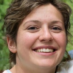 Bu, yüzünde parlak bir gülümseme olan bir kadının yakın çekim görüntüsüdür. Omuz hizasında dalgalı kahverengi saçları var ve gözleri memnun bir ifadeyle hafifçe kapalı. Dudakları hafifçe ayrılmış ve sıcak ve davetkâr bir gülümseme şeklinde yukarı kıvrılmış. Yanakları mutluluktan hafifçe kızarmış ve yüzü güneş tarafından aydınlatılmış. Arka planın düz, açık renkli bir duvar olması dikkatin kadının yüz hatlarına odaklanmasına yardımcı oluyor. Kadın rahatlamış ve o anın neşesiyle dolu görünüyor. Başı hafifçe sola doğru eğilmiş ve çenesi sanki hayranlıkla yukarı bakıyormuş gibi hafifçe kalkmış.