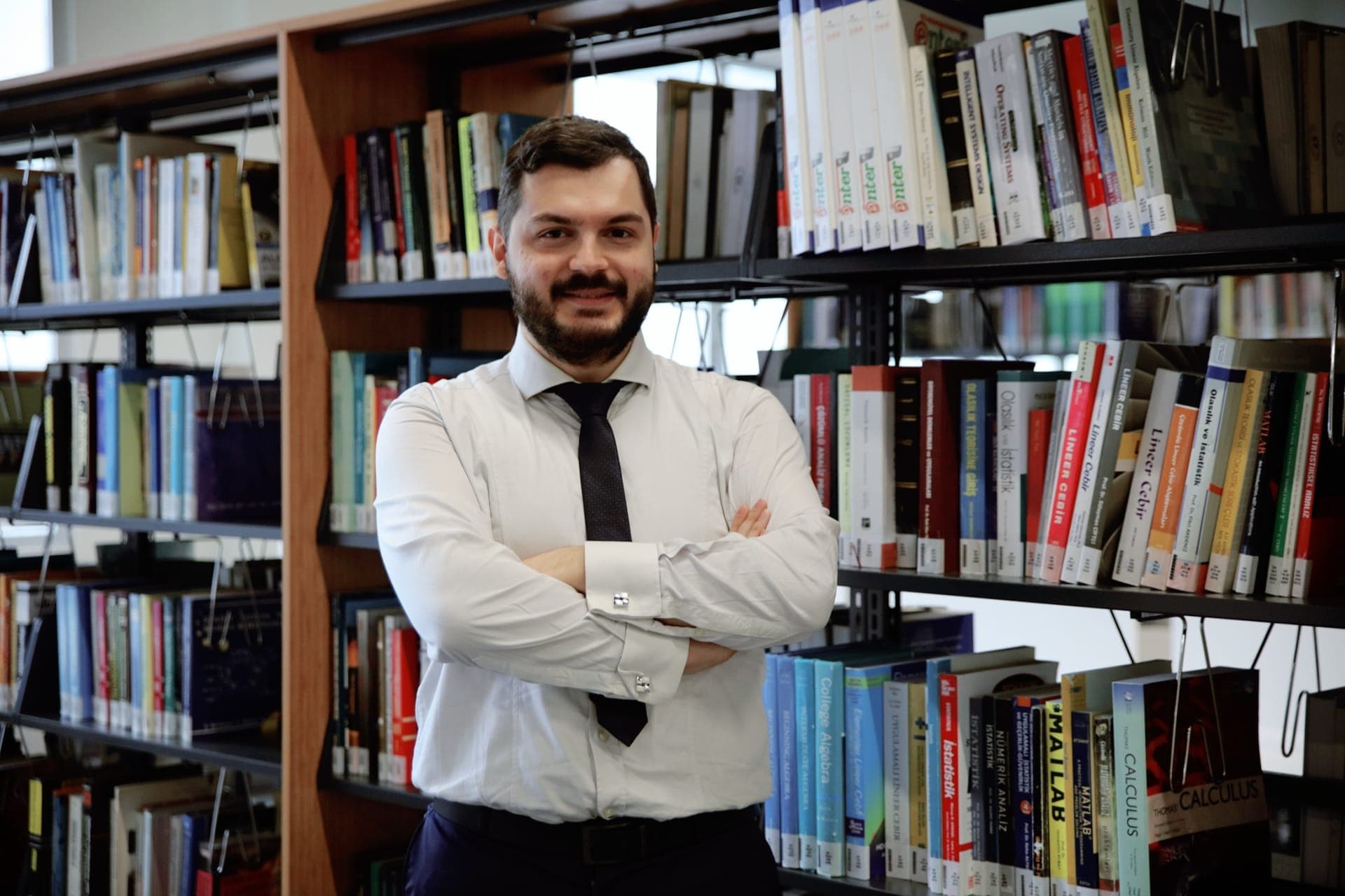 Bu fotoğrafta bir kitap rafının önünde duran bir eğitmen görülüyor. Sakalı var ve kravat takıyor. Eğitmen gülümsüyor, güven ve bilgi yayıyor. Yüzü yakın çekimle çerçevelenmiş ve yüz hatlarının detayları gösterilmiş. Daha yakından bakıldığında bir kol düğmesi ve bir kemer görülüyor. Arkasındaki kitaplar engin bilgisini ve entelektüel merakını simgeliyor. Raftaki bir kitabın yakın çekimi, eğitmenin öğrenme tutkusunu vurguluyor. Raftaki kitap sırası da konu hakkındaki uzmanlığını gösteriyor. asticaVision ai bu fotoğraftaki eğitmeni öğretme konusunda tutkulu ve alanında bilgili biri olarak tanıyabilir.