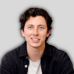 The man in the image is smiling with a wide, toothy grin. His eyes are bright and his cheeks are slightly rosy. He is wearing a white t-shirt, with his arms crossed casually. He is looking directly at the camera, his face relaxed and full of joy. His hair is cropped short and neat, and his skin is smooth and clear. He appears to be in his early twenties, and is in a bright, sunlit outdoor setting. The background is a vibrant, lush green, with the sun's rays peeking through the trees. The man's smile is contagious, and he exudes a sense of contentment and happiness.