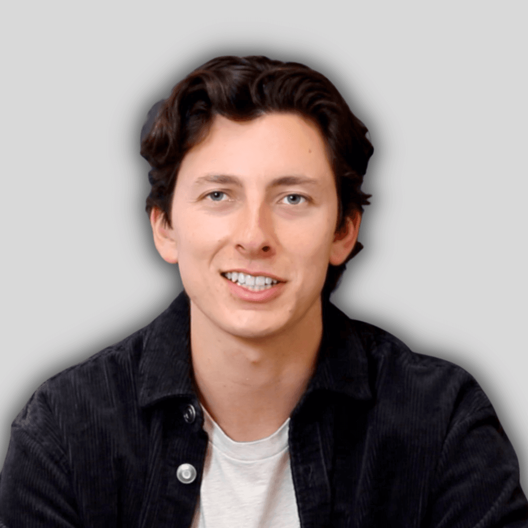 The man in the image is smiling with a wide, toothy grin. His eyes are bright and his cheeks are slightly rosy. He is wearing a white t-shirt, with his arms crossed casually. He is looking directly at the camera, his face relaxed and full of joy. His hair is cropped short and neat, and his skin is smooth and clear. He appears to be in his early twenties, and is in a bright, sunlit outdoor setting. The background is a vibrant, lush green, with the sun's rays peeking through the trees. The man's smile is contagious, and he exudes a sense of contentment and happiness.