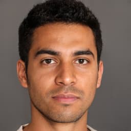 The man in the image has short, dark hair that is neatly groomed and styled. His face is oval in shape with a strong jawline and a short, straight nose. His eyes are a dark brown color and he has thick eyebrows. His lips are full and curved and he has a confident expression on his face. He is wearing a collared shirt with the top button undone and no tie. His outfit is completed with a pair of dark-colored pants and black dress shoes. He stands with his arms crossed and his posture is confident and strong.