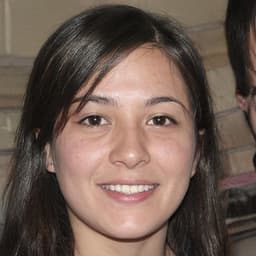 A young woman with long brown hair is smiling widely at the camera. She has a bright and friendly expression on her face, her eyes crinkled in joy. She is wearing a white t-shirt with a bright pink scarf around her neck. Her arms are relaxed and she is standing in a bright and sunny outdoor environment, with lush green trees in the background. She looks confident and content, her smile radiating warmth and happiness. Her cheeks are rosy and her lips are slightly parted, conveying a sense of excitement. This image is a perfect representation of joy and contentment.
