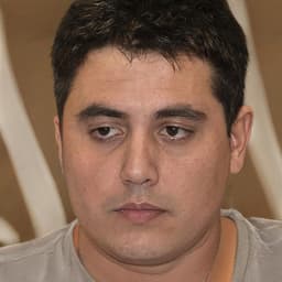 The man is looking down with a serious expression on his face. He is wearing a black t-shirt with grey trousers and white sneakers. His eyes are dark and his hair is short and dark. His arms are crossed in front of him and his body is slightly hunched over. He has a stern and determined look on his face with a slight frown. His lips are slightly parted and his jaw is set. His brows are furrowed and his eyebrows are slightly raised. He looks deep in thought, lost in his own thoughts and contemplative. The background is a plain white wall with a few posters and decorations. He looks thoughtful and introspective, as if he is reflecting deeply on something.
