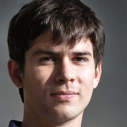 A man with short brown hair sits in front of a white background. He is wearing a navy blue t-shirt and dark blue jeans. His facial expression is serious and he has a determined look on his face. His hair is neatly combed and styled in a short, side swept style. His eyes are brown and his eyebrows are arched in concentration. His arms are resting on his knees and his hands are clasped together in front of him. He looks like he is deep in thought, trying to figure out a difficult problem. His posture is confident and he looks like he is ready to tackle any challenge that comes his way.