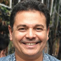 The man is standing in front of a white background, wearing a dark shirt and jeans. He has a big smile on his face, showing off his perfect white teeth. His eyes are closed, accentuating his happiness. His hair is tousled and he has a small dimple on his cheek. He looks relaxed and content, thrilled to be having his picture taken. His arms are crossed in front of him, and his fingers are lightly intertwined. He looks to be in his late twenties, with a youthful energy that is contagious. He is the embodiment of joy, radiating a true sense of inner peace.