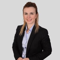 Bu, profesyonel iş kıyafeti giyen genç bir kadının fotoğrafı. Mavi duvarlı ve kahverengi halılı iyi aydınlatılmış bir odada duruyor. Kadının elleri cebinde ve duruşu kendinden emin ve dengeli. Koyu renk saçları düzgün bir şekilde toplanmış ve yüzü hafifçe makyajlanmış. Lacivert bir takım elbise ceketi, altında beyaz bir bluz ve lacivert bir pantolon giyiyor. Siyah ayakkabıları cilalı ve ona sofistike bir görünüm veriyor. Gururlu ve kendinden emin görünüyor ve ifadesi sakin ve odaklanmış.
