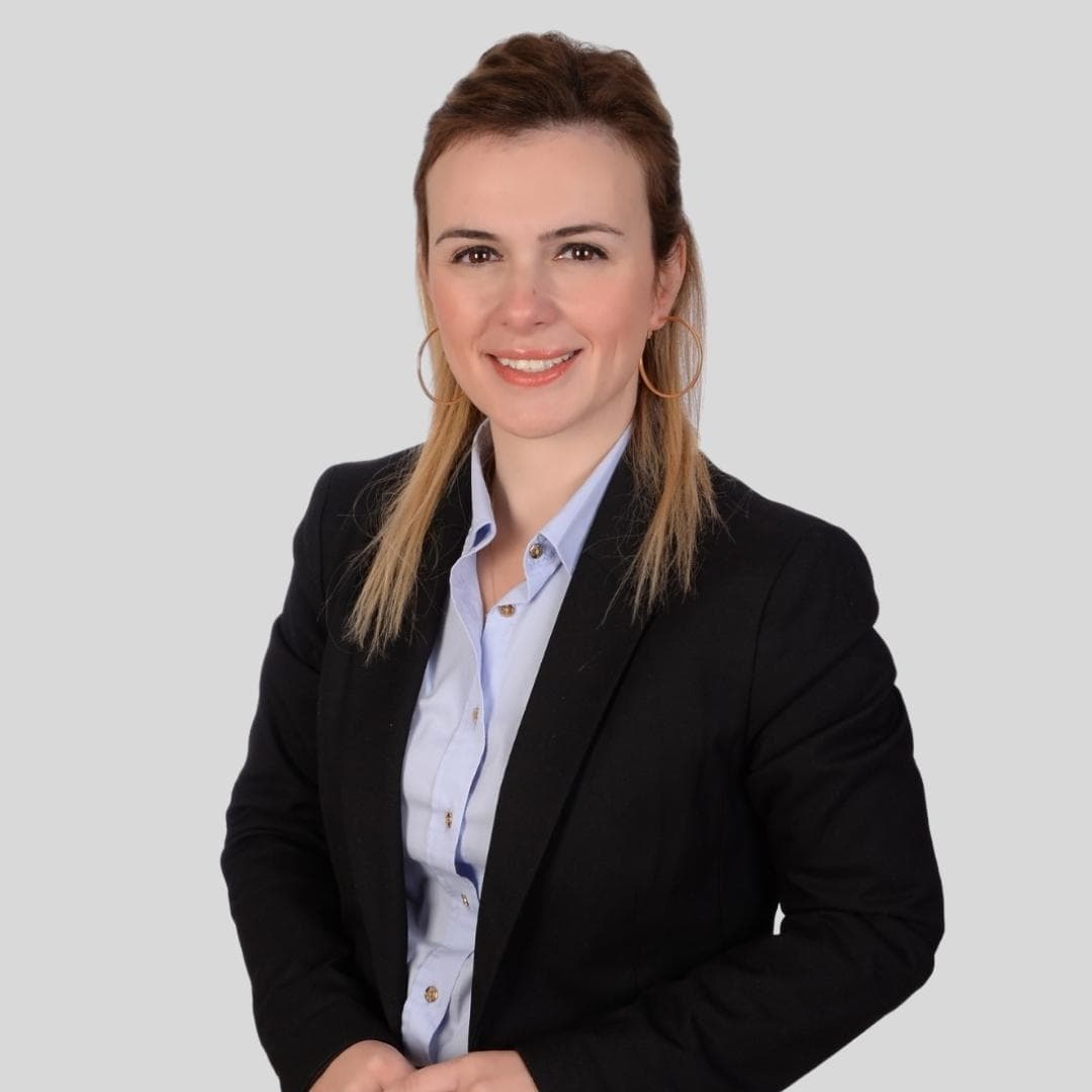 This is a photo of a young woman wearing a professional business suit. She is standing in a well-lit room with a blue wall and brown carpet. The woman has her hands in her pockets, and her posture is confident and poised. Her dark hair is styled in a neat updo, and her face is lightly made-up. She is wearing a navy blue suit jacket with a white blouse underneath, and navy blue trousers. Her black shoes are polished and give her a look of sophistication. She looks proud and self-assured, and her expression is calm and focused.