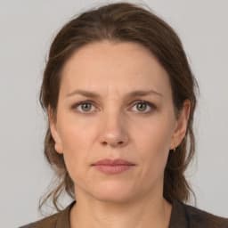 This close-up image of a woman is softly lit and her features can be made out clearly. She has a heart shaped face with high cheekbones and a pointed chin. Her eyes are a warm hazel color and her lips are slightly parted. Her hair is a dark brown color and falls just past her shoulders. She is wearing a white t-shirt and her hands are clasped in front of her. The overall tone of the image is peaceful and warm. She looks to be in a calm and relaxed state, looking off into the distance with a thoughtful expression.
