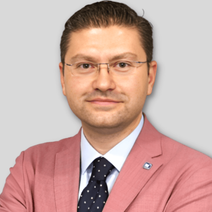 A handsome, well-dressed man wearing a tailored black suit and necktie stands tall and confident. He has a light-colored button-down shirt beneath his suit, and his strong jawline is emphasized by his neatly groomed facial hair. His posture is relaxed and he stands with his hands in his pockets, giving him an air of self-assurance. His dark hair is styled in a comb-over, and his eyes stare directly ahead with a determined gaze.
