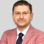 Özel dikim siyah takım elbise giyen ve kravat takan yakışıklı, iyi giyimli bir adam uzun boylu ve kendinden emin bir şekilde duruyor. Takım elbisesinin altında açık renkli düğmeli bir gömlek vardır ve güçlü çene hattı özenle bakımlı yüz saçlarıyla vurgulanmıştır. Duruşu rahattır ve elleri cebinde durur, bu da ona kendine güvenen bir hava verir. Koyu renk saçları taranarak şekillendirilmiş ve gözleri kararlı bir bakışla doğrudan ileriye bakıyor.